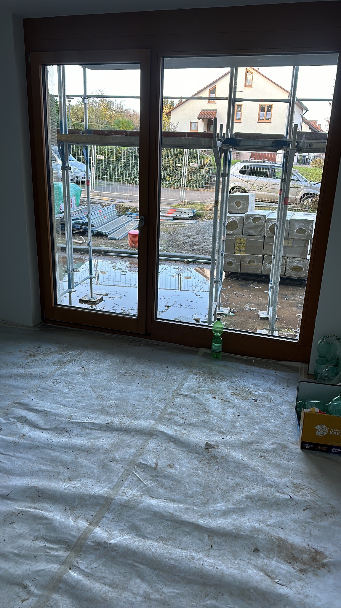 Montaż okien balkonowych z brązową ramą w surowym stanie. Na zewnątrz widoczne rusztowania, materiały budowlane i dom.