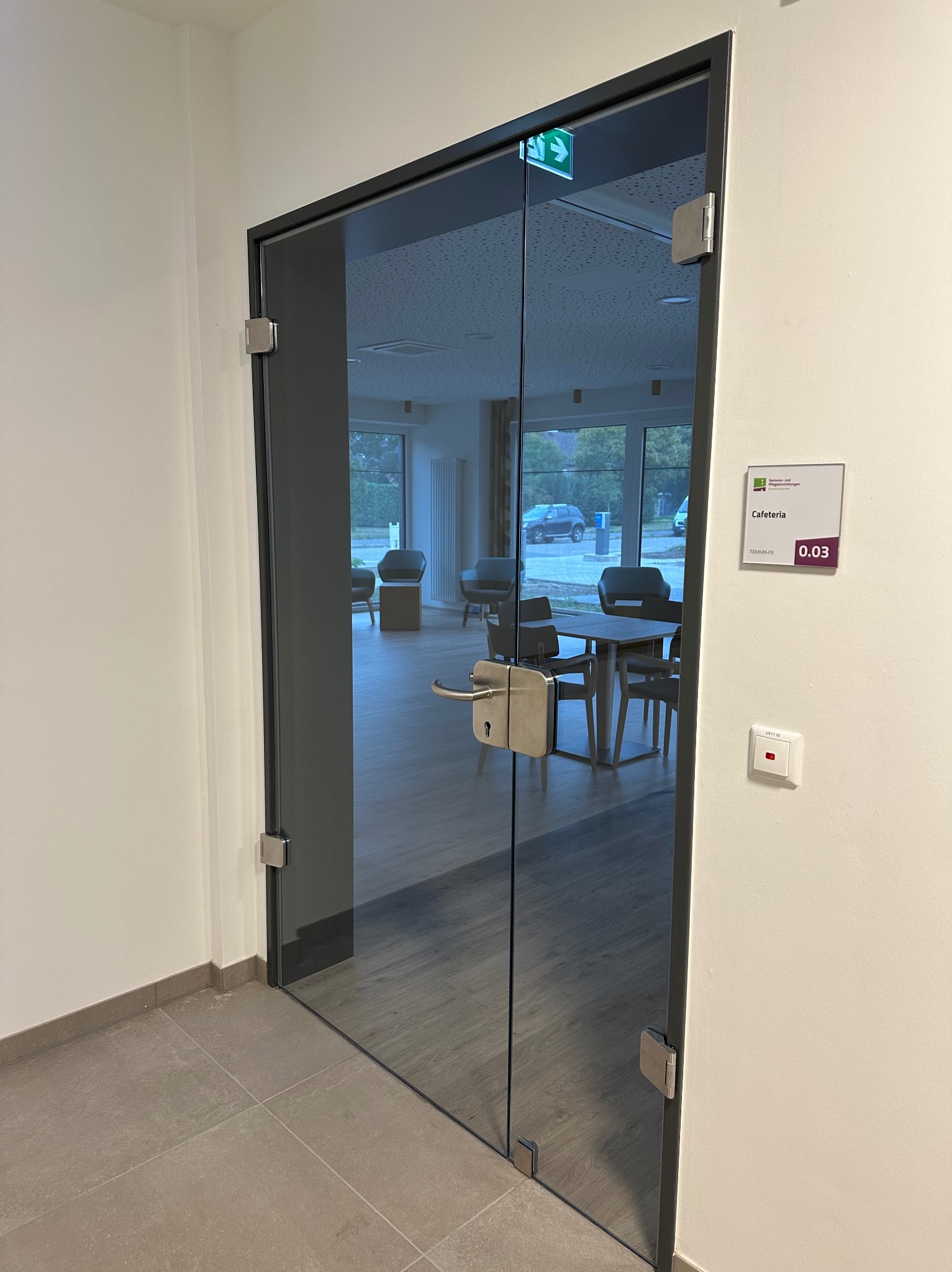 Szklane drzwi z metalową ramą, prowadzące do kawiarni z widocznym wnętrzem i oznaczeniem numeru pokoju 0.03, oświetlone światłem dziennym.