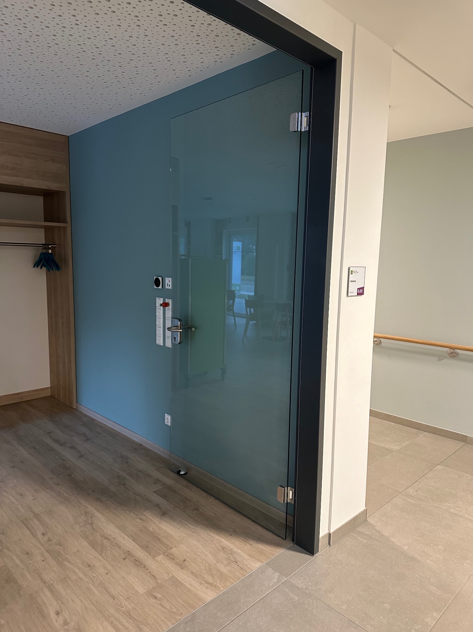 Szklane drzwi z czarną ramą, zamek elektroniczny, widok na wnętrze. Podłoga z paneli, błękitna ściana. Widoczna klamka i zawiasy. Minimalistyczny design.