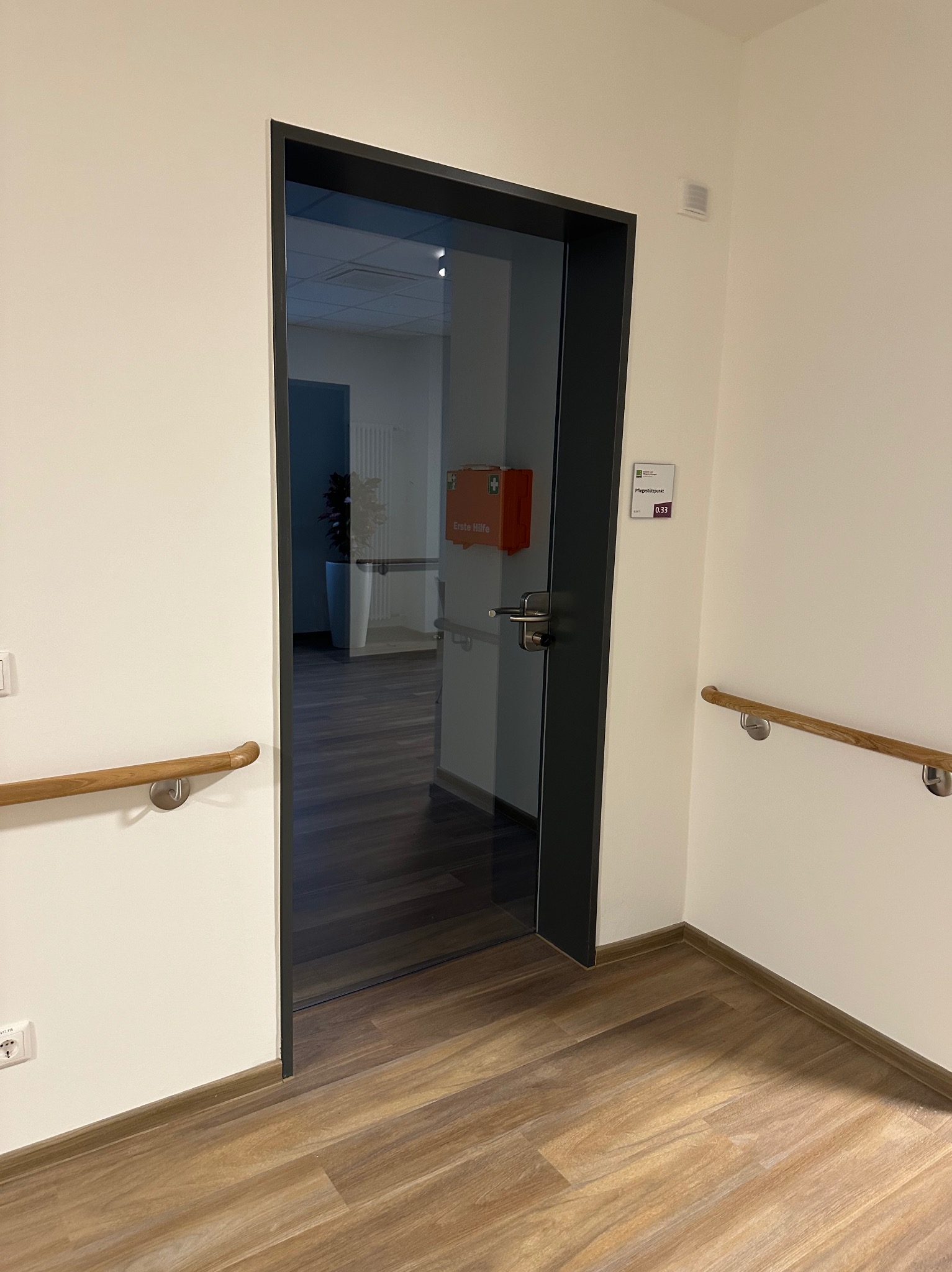 Szklane drzwi wewnętrzne z czarną ramą w jasnym pomieszczeniu, widoczna apteczka pierwszej pomocy w tle. Podłoga z paneli imitujących drewno.