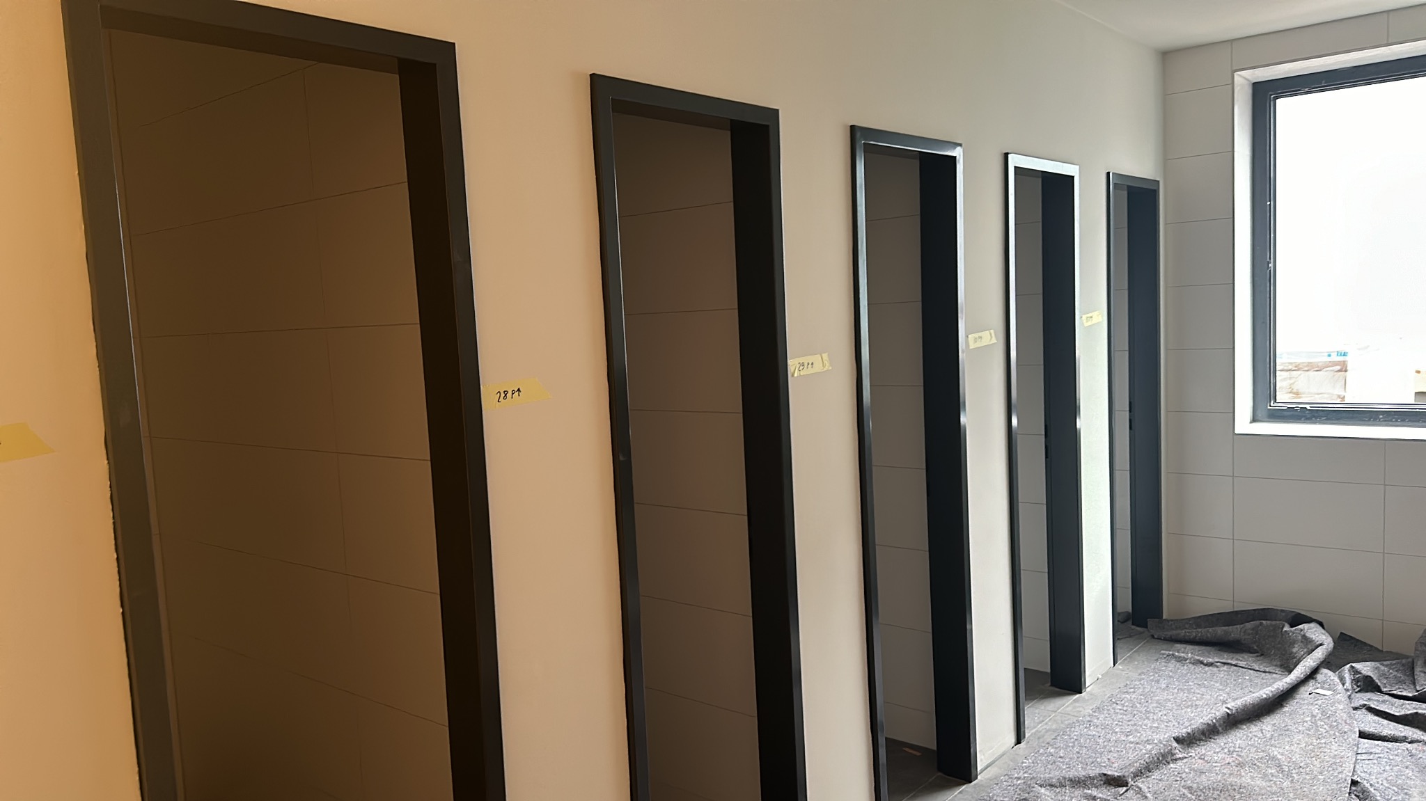 Rząd zamontowanych, nowoczesnych drzwi wewnętrznych z czarnymi ościeżnicami w jasnym pomieszczeniu z wykończeniem w płytkach. Na drzwiach widoczne żółte karteczki.