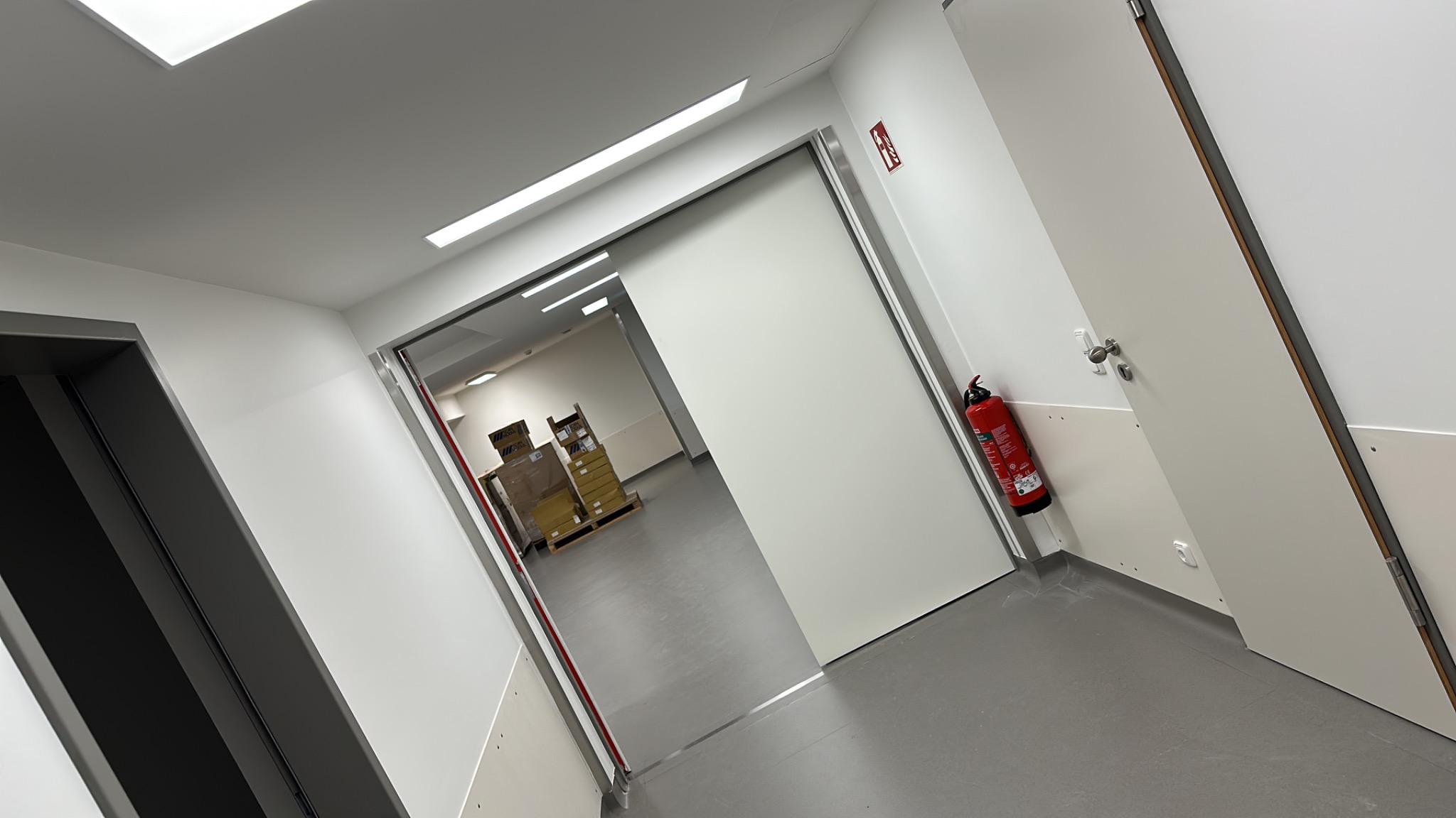 Nowoczesne, przesuwne drzwi wewnętrzne z czerwonym znakiem ewakuacyjnym i gaśnicą w tle, w sterylnym, białym korytarzu, ujęcie pod kątem.