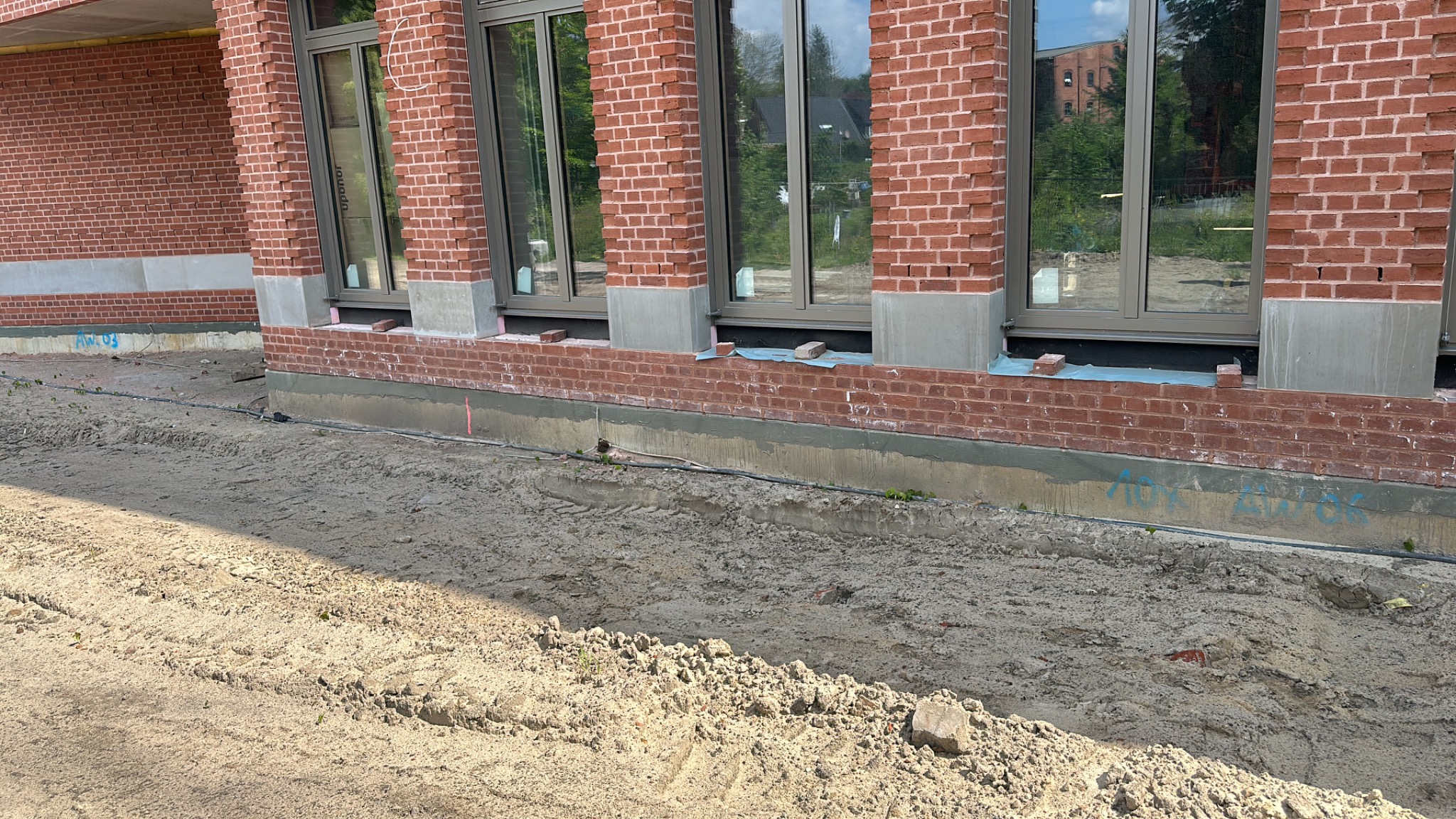 Budynek z cegły z zamontowanymi oknami z szarymi ramami, widoczne przygotowania do dalszych prac wykończeniowych wokół budynku.