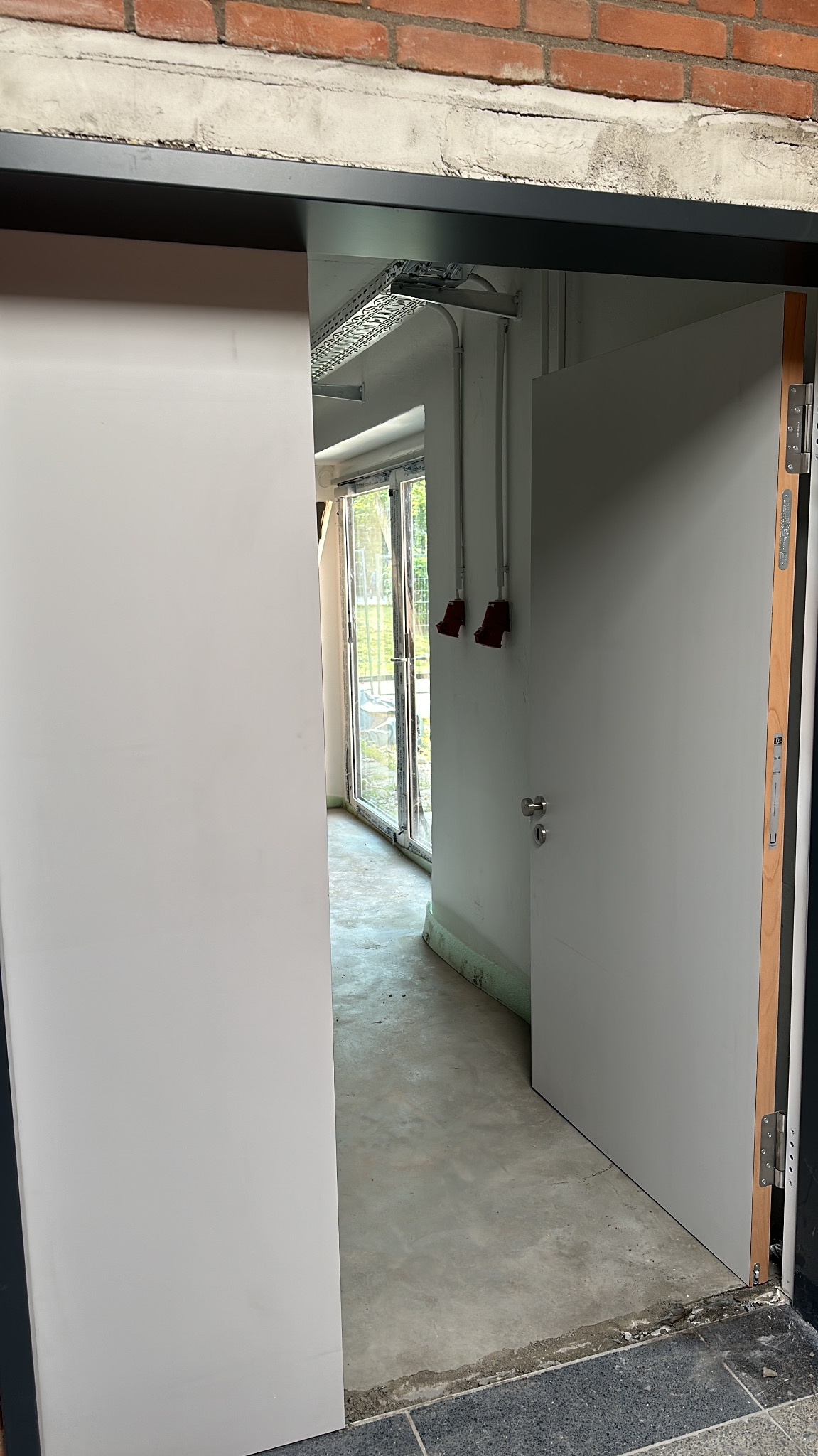 Wnętrze z widokiem na przesuwne, białe drzwi i okno. Widoczne są elementy instalacji elektrycznej i surowy beton na podłodze. Ceglany fragment ściany w górnej części kadru.