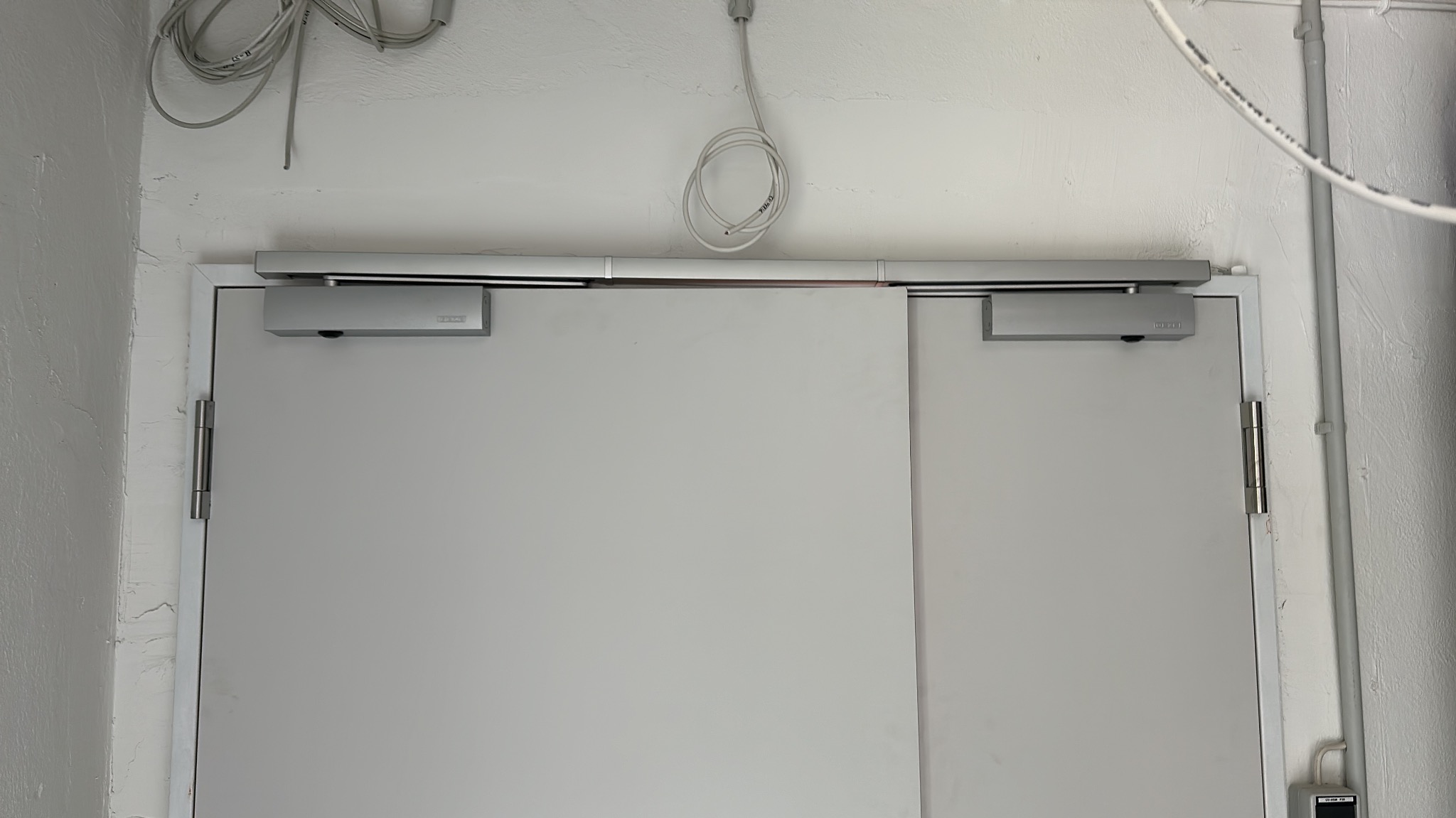 Białe, dwuskrzydłowe drzwi z samozamykaczami Dorma, zamontowane w białej futrynie na tle białej ściany z widoczną instalacją elektryczną.