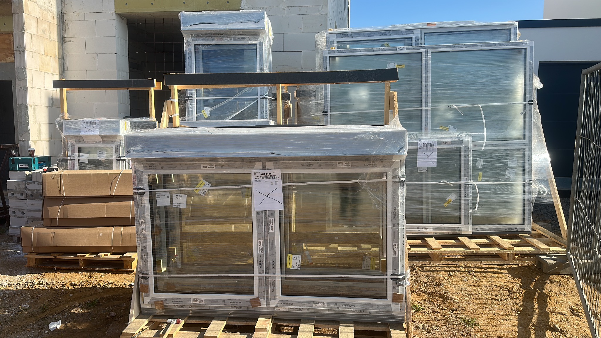 Nowe okna PCV Salamander, zabezpieczone folią, ułożone na paletach na placu budowy. Widoczne cegły i narzędzia. Słoneczny dzień.