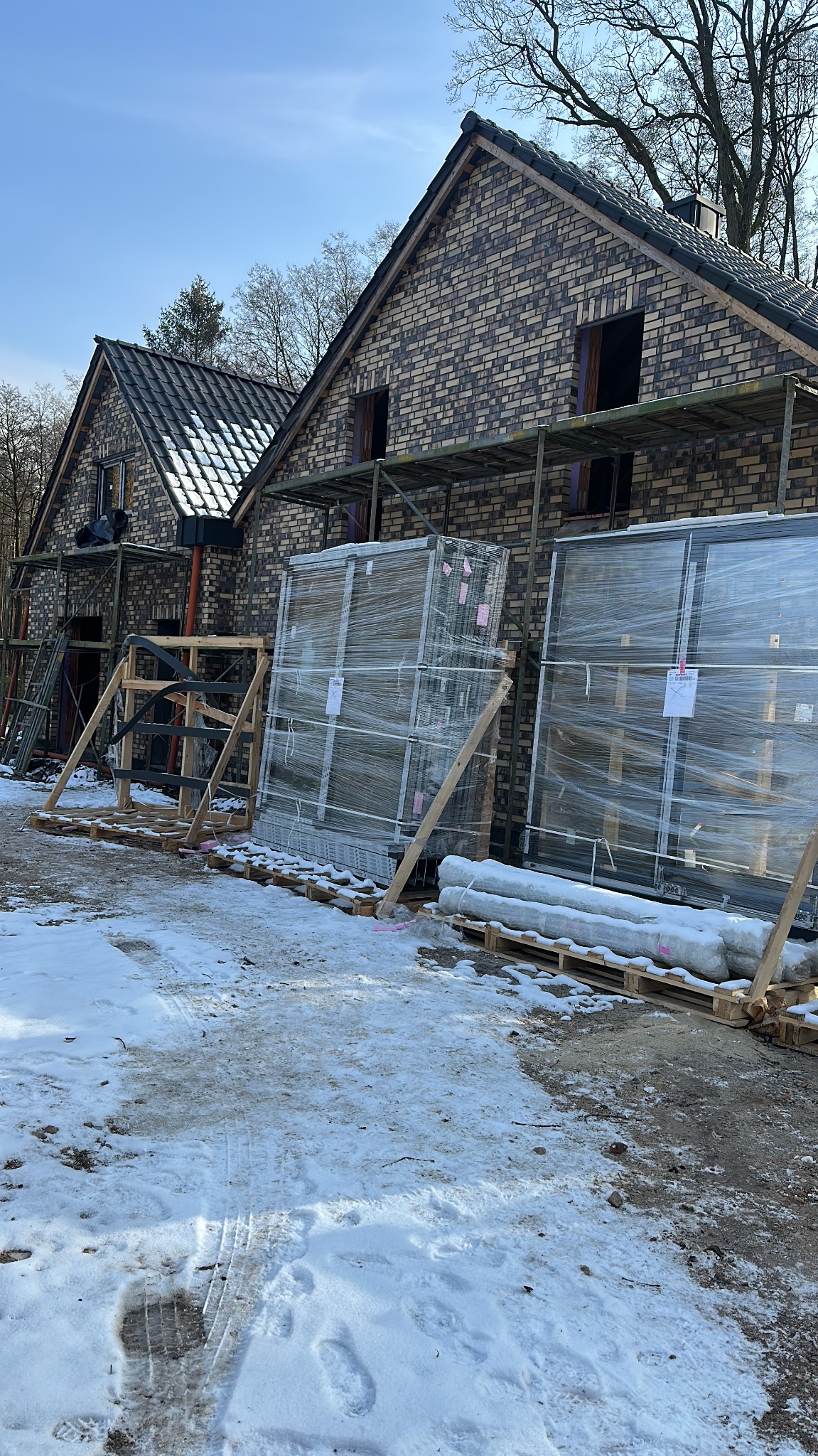 Nowe okna PCV w folii na paletach, czekają na montaż przy domu z cegły w zimowej scenerii. Widoczne rusztowania i przygotowania do instalacji.