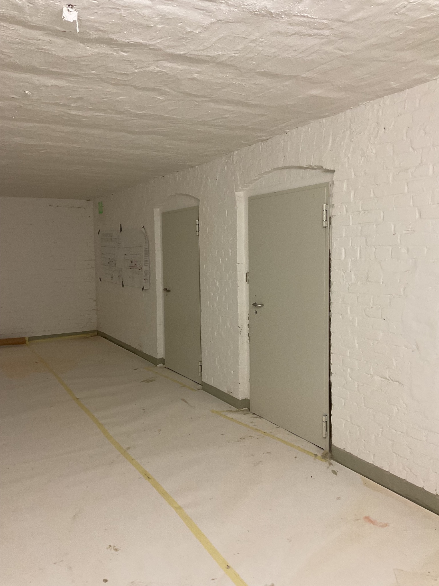 Wnętrze piwnicy z dwiema szarymi, metalowymi drzwiami i białymi, pomalowanymi ceglanymi ścianami. Podłoga zabezpieczona folią i taśmą malarską, widoczny plan na ścianie.