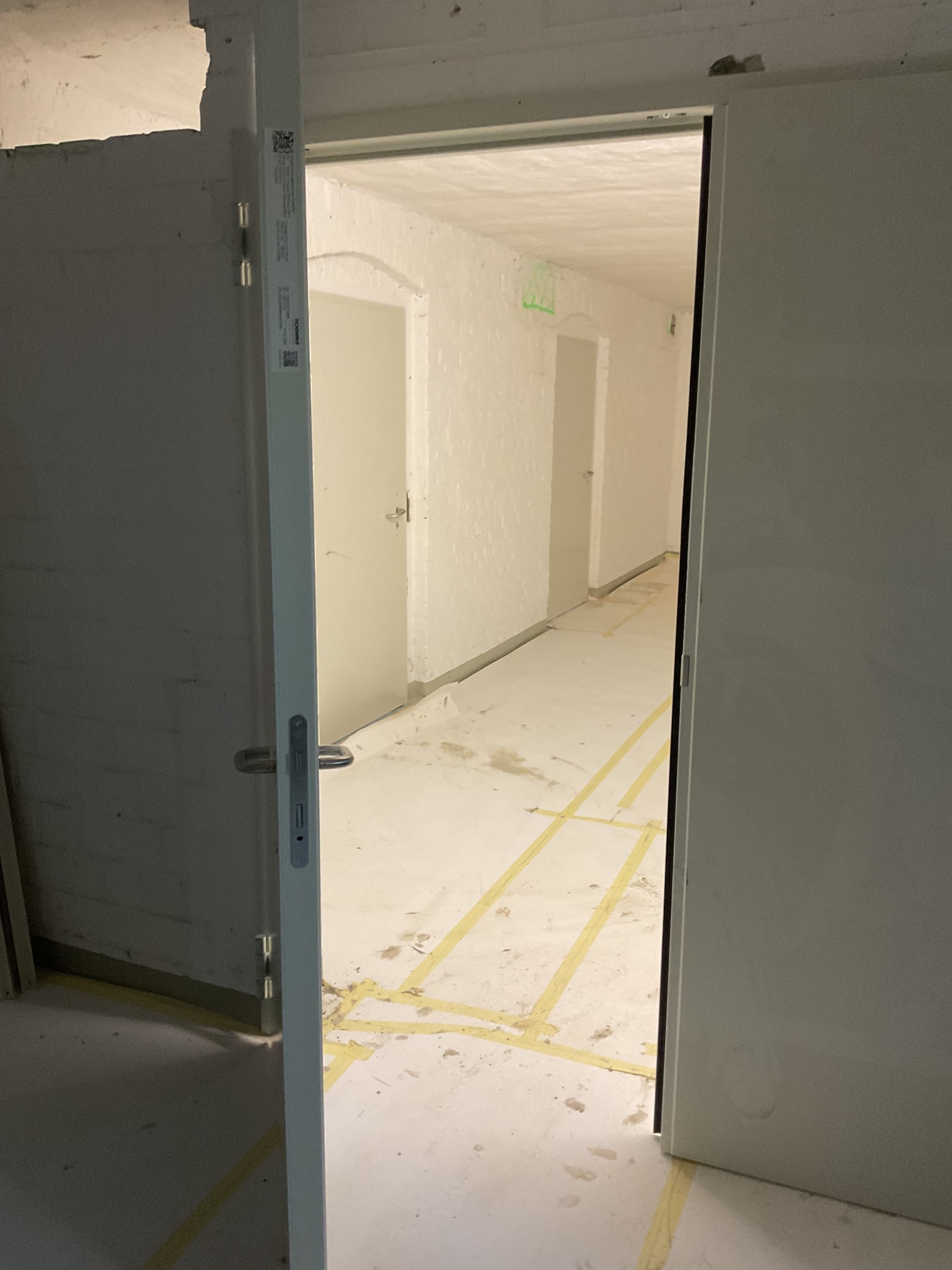 Otwarta, biała, wewnętrzna ościeżnica z widokiem na korytarz z białymi drzwiami i surowymi ścianami. Żółta taśma na podłodze wyznacza strefy.