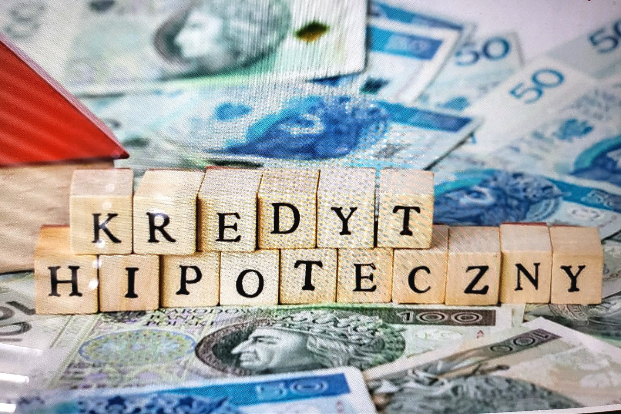 Drewniane klocki z napisem 'KREDYT HIPOTECZNY' na tle banknotów polskich złotych i miniatury domu z czerwonym dachem.