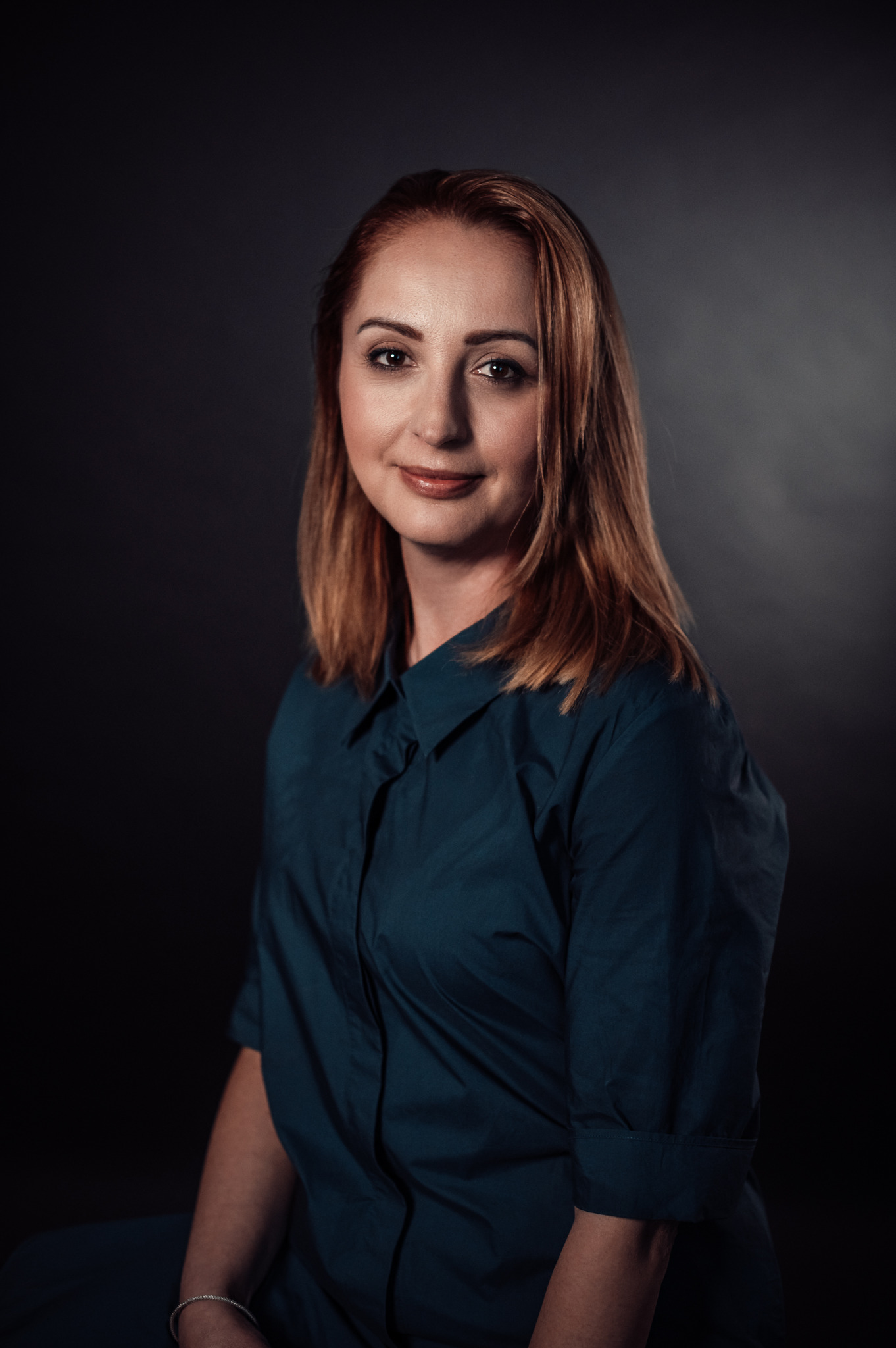 Portret kobiety z rudymi włosami, ubrana w ciemnozieloną koszulę, uśmiechająca się na ciemnym tle.