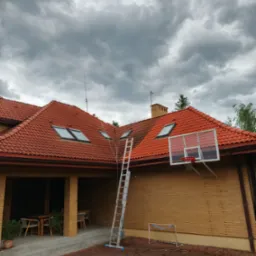Częściowo umyty, czerwony dach z oknami dachowymi, drabina oparta o dach, kosz do koszykówki na ścianie budynku. Pochmurne niebo w tle.