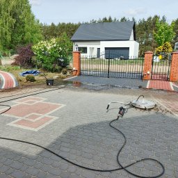 Grzegorz Modrzejewski - Mycie kostki brukowej na posesji. Widoczna myjka ciśnieniowa z dyszą rotacyjną i wąż. Część kostki umyta, ukazując różnicę w kolorze. Dom i brama w tle.