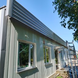 Grzegorz Modrzejewski - Szara elewacja budynku z blachy trapezowej po czyszczeniu, z odbiciami w oknach i drabiną opartą o ścianę. Widoczne ślady wody po myciu.