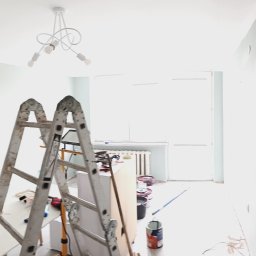 Grzegorz Modrzejewski - Pokój w trakcie malowania na pastelowy odcień, widoczne drabiny malarskie, pędzle i farby. Ściany częściowo pomalowane, sufit biały, w tle okno i grzejnik.