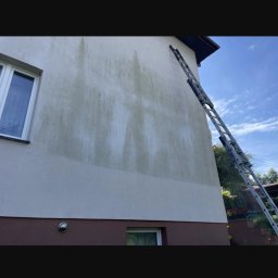 Fragment elewacji budynku z widocznymi śladami czyszczenia, z drabiną opartą o ścianę. Częściowo umyta elewacja kontrastuje z zabrudzoną powierzchnią.