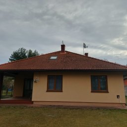 Grzegorz Modrzejewski - Ujęcie domu z beżową elewacją i czerwoną dachówką. Widoczny fragment ogrodu, boczna elewacja z oknami i zadaszony taras z ceglaną kolumną.