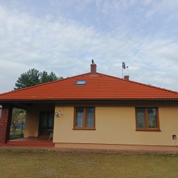 Grzegorz Modrzejewski - Dom jednorodzinny z czerwoną dachówką, beżową elewacją i widoczną instalacją satelitarną. Na kominie dekoracja w kształcie orła. Wiosenne drzewa w tle.