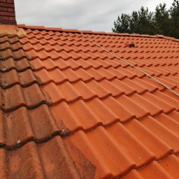 Grzegorz Modrzejewski - Częściowo wyczyszczony dach z czerwonej dachówki, widoczny komin z cegły i lina zabezpieczająca. Po lewej stronie dach pokryty mchem i zabrudzeniami, po prawej czysty.