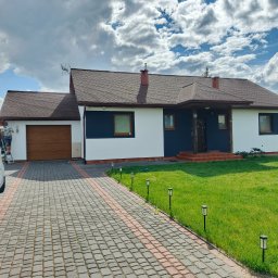 GrzesiekMaluje - Dwukondygnacyjny dom z pomalowaną elewacją w tonacji bieli i granatu. Na podjeździe stoi samochód dostawczy, a obok niego pracownik na drabinie.