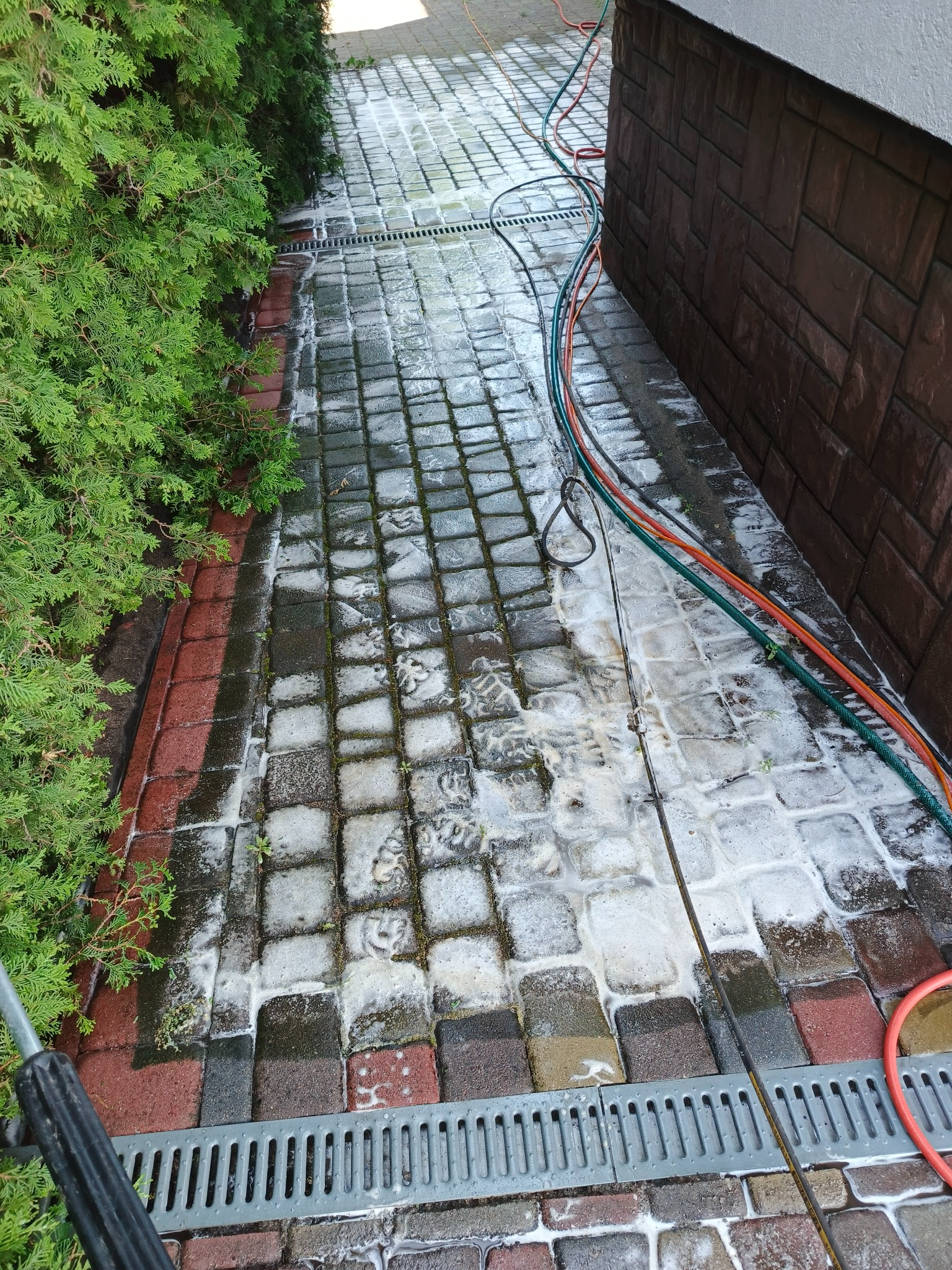Fragment mytej kostki brukowej z widoczną pianą i dyszą myjki ciśnieniowej, w tle zielony żywopłot i elewacja budynku. Wzdłuż kostki biegną węże ogrodowe.