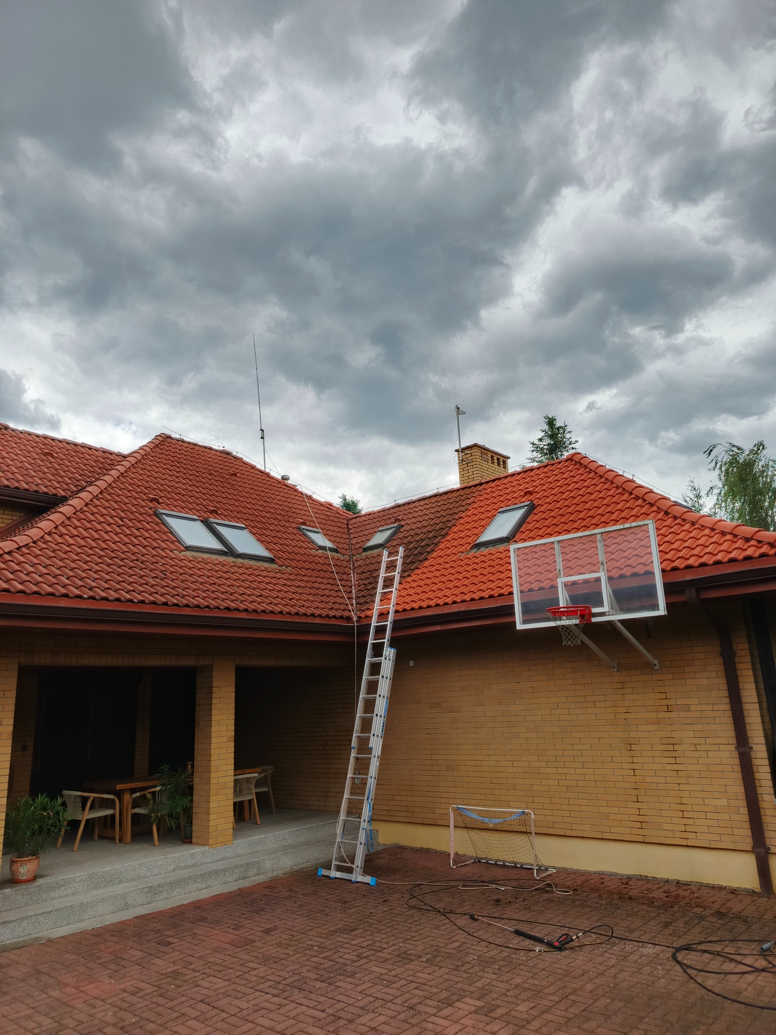 Częściowo umyty, czerwony dach z oknami dachowymi, drabina oparta o dach, kosz do koszykówki na ścianie budynku. Pochmurne niebo w tle.