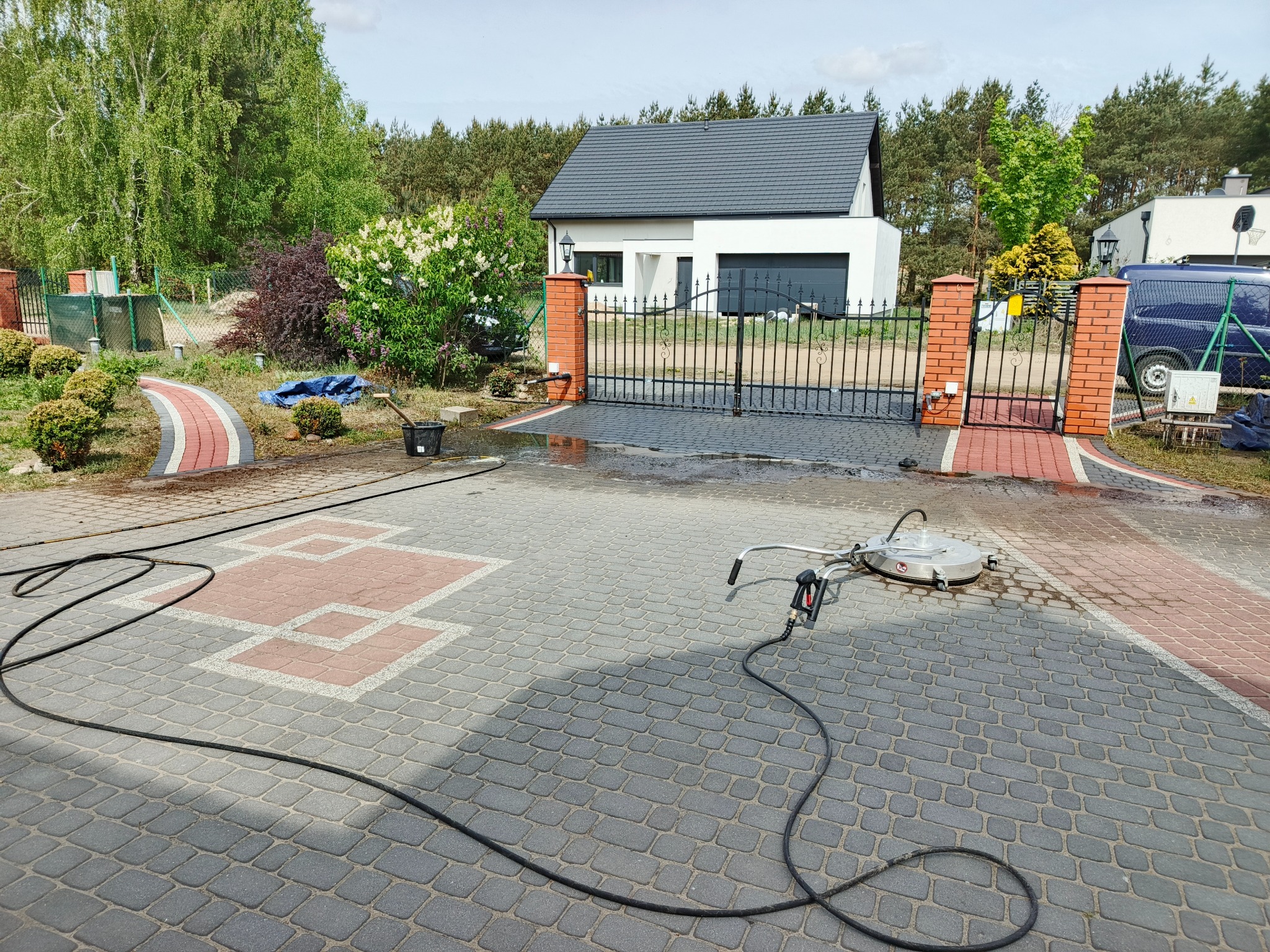 Mycie kostki brukowej na posesji. Widoczna myjka ciśnieniowa z dyszą rotacyjną i wąż. Część kostki umyta, ukazując różnicę w kolorze. Dom i brama w tle.