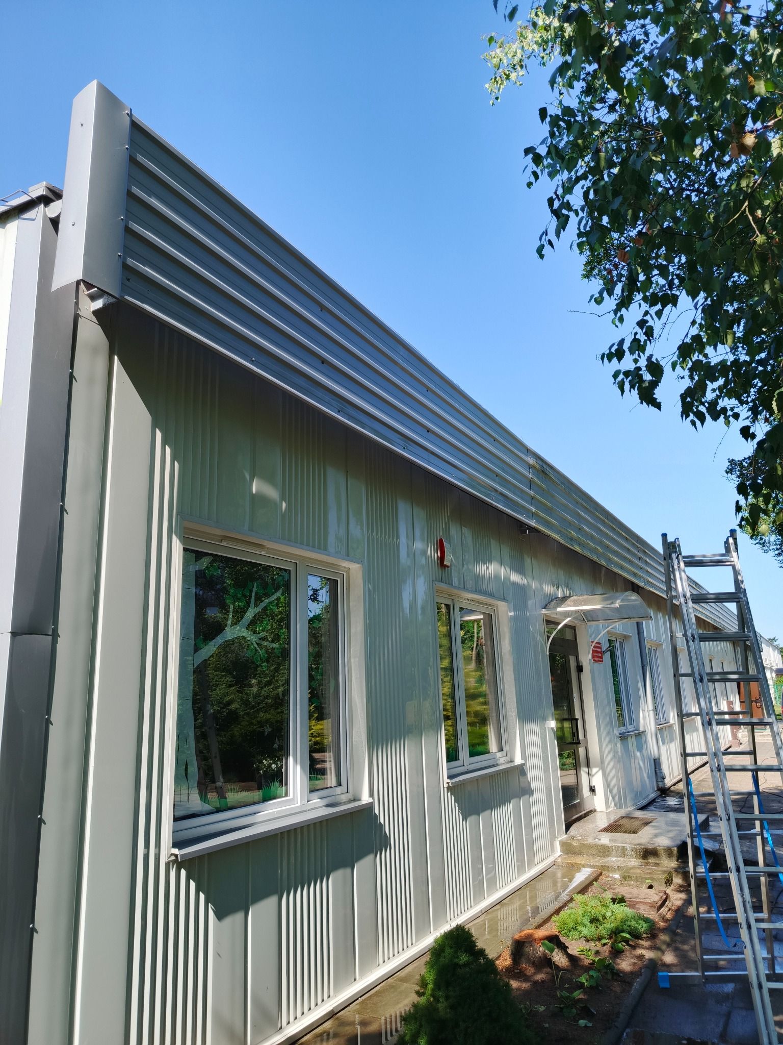 Szara elewacja budynku z blachy trapezowej po czyszczeniu, z odbiciami w oknach i drabiną opartą o ścianę. Widoczne ślady wody po myciu.