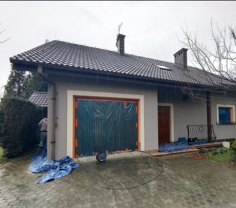 Przygotowanie do malowania elewacji domu: zabezpieczona folią brama garażowa, okno i drzwi wejściowe. Widoczny fragment elewacji w szarym kolorze i narzędzia.