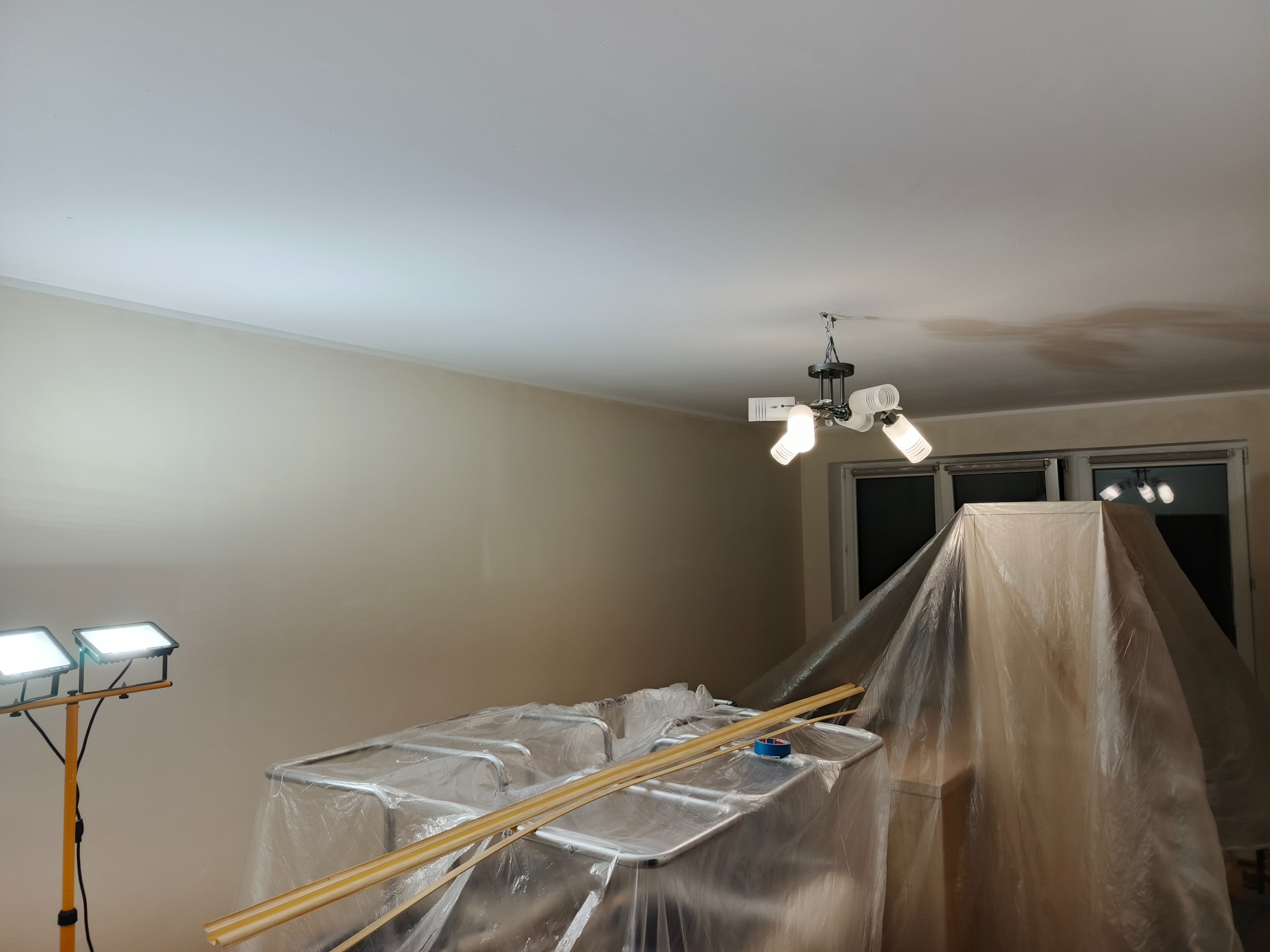 Przygotowania do malowania: meble zabezpieczone folią, listwy przypodłogowe, oświetlenie robocze. Ściany w odcieniach beżu, sufit biały z widoczną lampą.