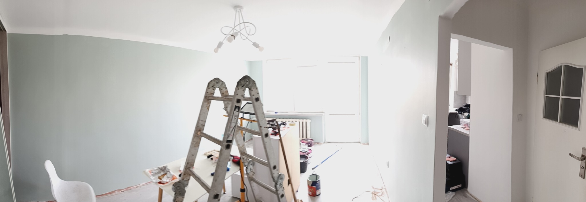 Pokój w trakcie malowania na pastelowy odcień, widoczne drabiny malarskie, pędzle i farby. Ściany częściowo pomalowane, sufit biały, w tle okno i grzejnik.