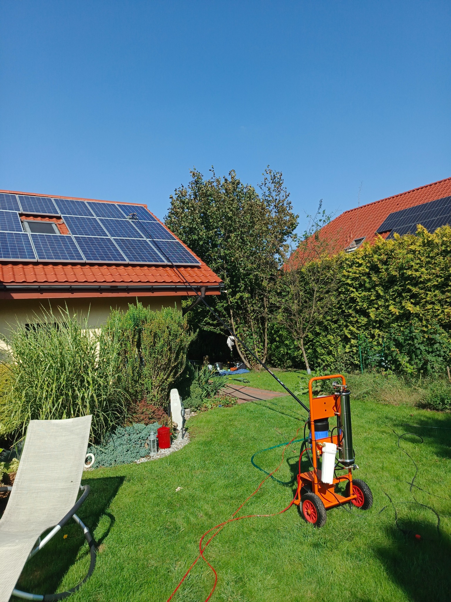 Pomarańczowy wózek do mycia paneli fotowoltaicznych na tle zielonego trawnika i domu z panelami słonecznymi w słoneczny dzień. Widoczny wąż i lanca teleskopowa.