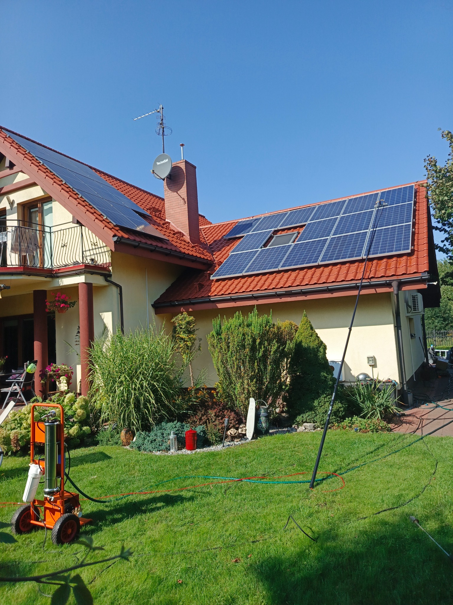 Dom z panelami słonecznymi czyszczonymi za pomocą specjalistycznego sprzętu. W tle błękitne niebo i zadbany ogród. Widoczny wąż i urządzenie do mycia.