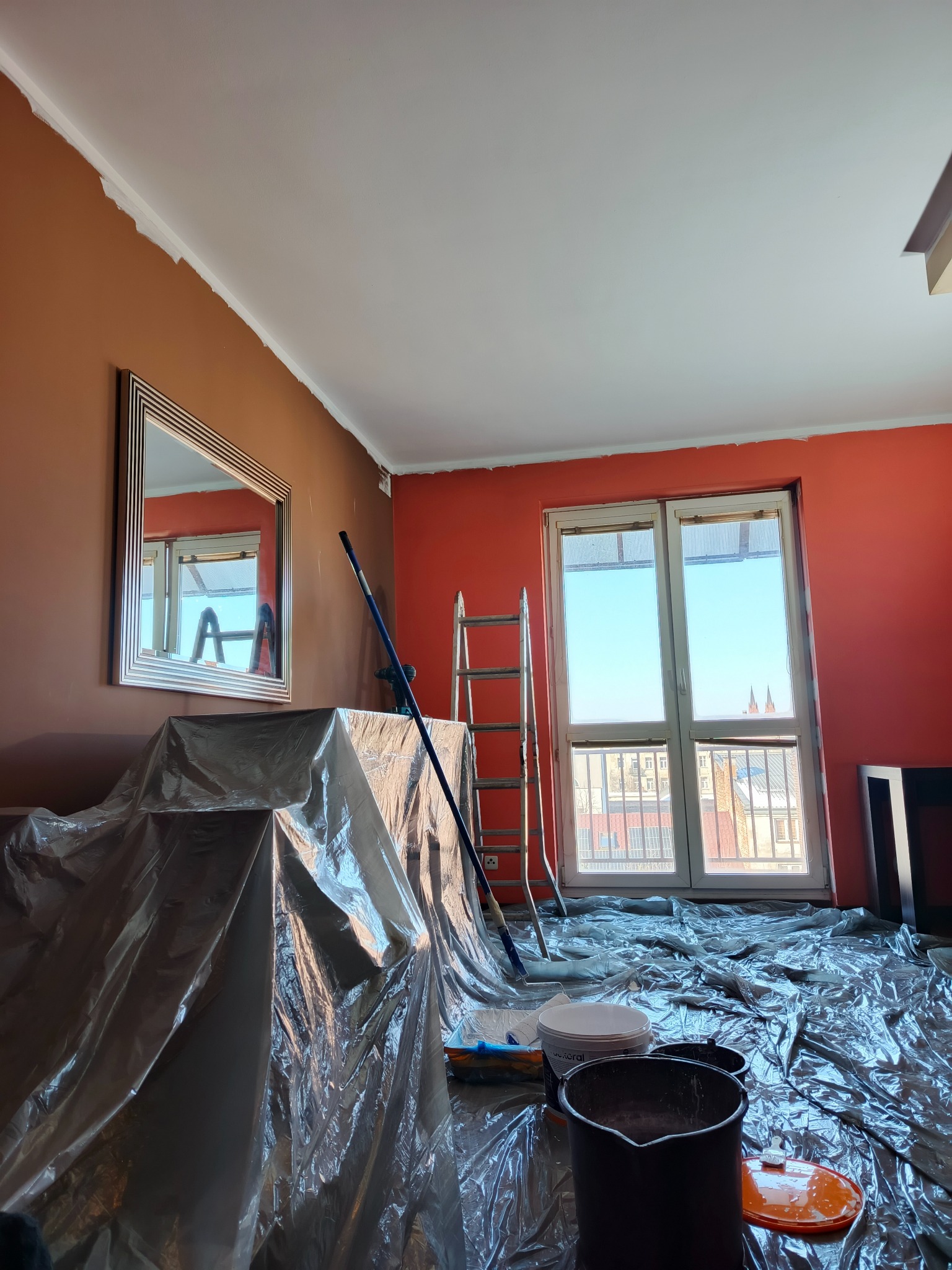 Pokój w trakcie malowania na odcienie czerwieni i brązu, zabezpieczony folią malarską. Widoczne drabina, wałek i pędzle. W tle okno z widokiem na miasto.