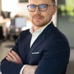 Portret mężczyzny w okularach i granatowej marynarce, z założonymi rękami, w jasnym biurze. Uśmiech, kr&oacute;tka broda, nowoczesne wnętrze w tle.