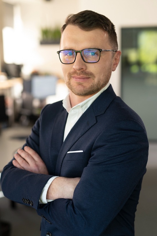 Portret mężczyzny w okularach i granatowej marynarce, z założonymi rękami, w jasnym biurze. Uśmiech, krótka broda, nowoczesne wnętrze w tle.