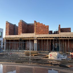 Budowa domu z czerwonej cegły: wzniesione ściany parteru, widoczne podpory i materiały budowlane. Stan surowy otwarty w słoneczny dzień.