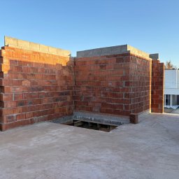 Wznoszone ściany z czerwonej cegły na fundamencie budynku w trakcie budowy. Widoczny otwór w podłodze i fragment budynku w tle, w słoneczny dzień.