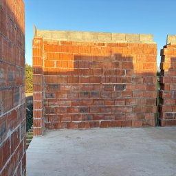 Wznoszone ściany z czerwonej cegły ceramicznej na fundamencie, widoczne spoiny i pustaki. Budowa domu w trakcie realizacji, w tle błękitne niebo i zieleń drzew.