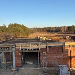 JULAS SOLUTIONS Julia Machnicka - Budowa domu z czerwonej cegły: ściany, szalunki, zbrojenie stropu. Widok z góry na plac budowy z lasem w tle, w słoneczny dzień.