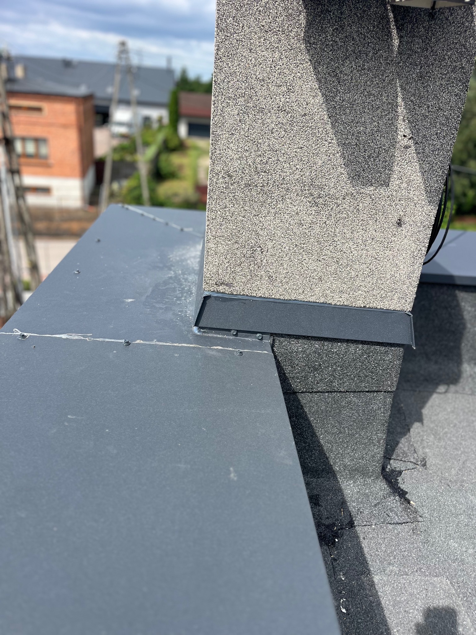 Szczegółowe ujęcie dachu z papy z kominem wykończonym szarym tynkiem strukturalnym, na tle zabudowy jednorodzinnej i błękitnego nieba.