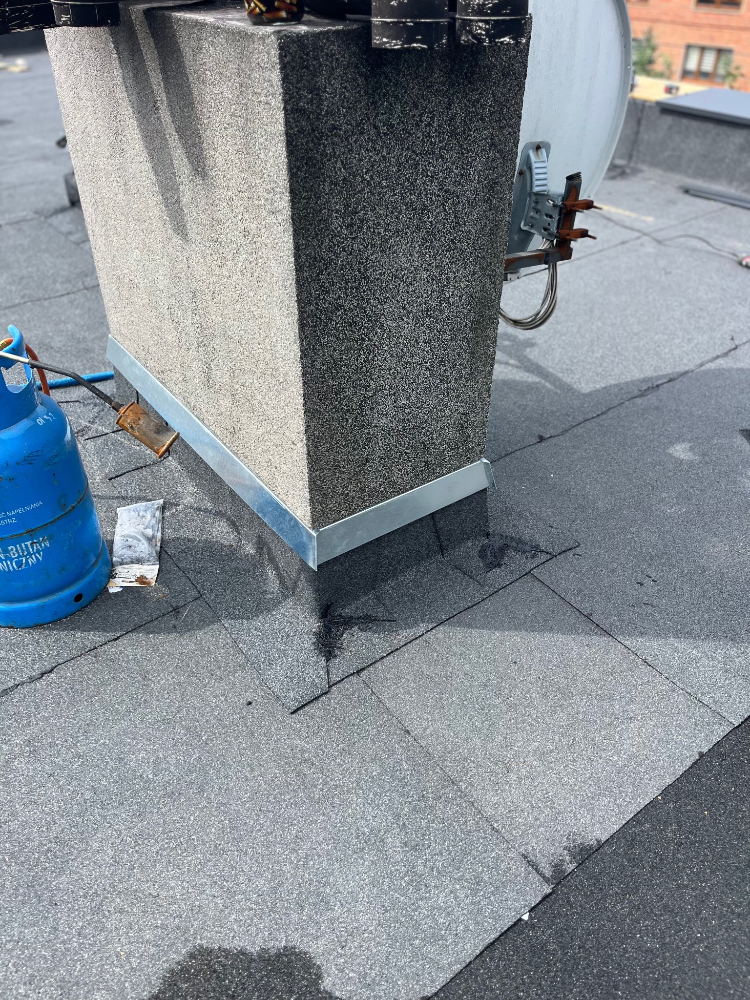 Szary komin z metalowym obramowaniem na pokrytym papą dachu. Widoczna butla gazowa i narzędzia dekarskie. Ujęcie z góry na fragment dachu.