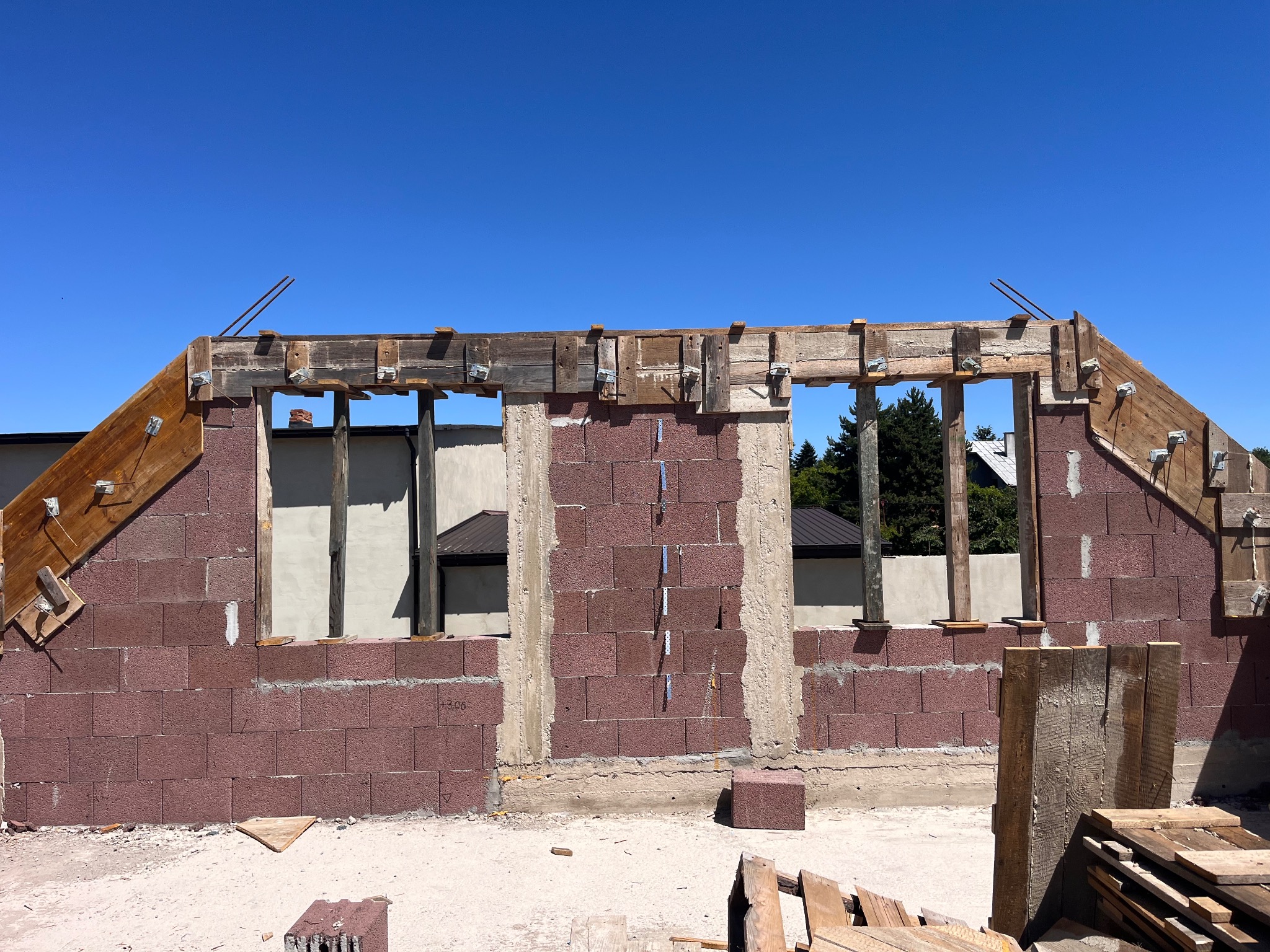 Budowa domu z czerwonych bloczków, widoczne drewniane szalunki okienne i zbrojenie dachu, błękitne niebo w tle. Etap wznoszenia ścian.