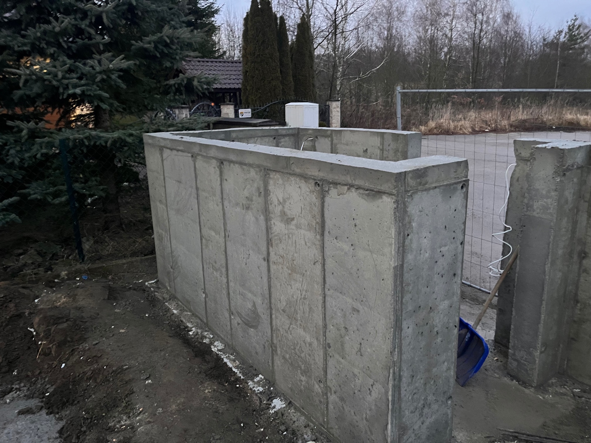 Betonowa konstrukcja fundamentowa w trakcie budowy domu, widoczne szalunki i teren budowy. W tle dom z ciemnym dachem i zielone drzewa.