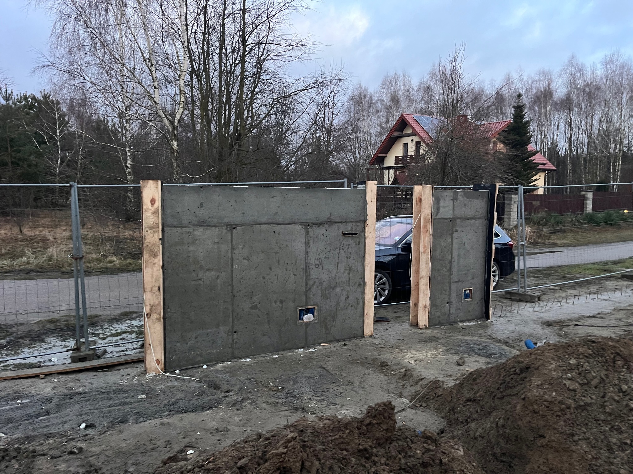Surowy betonowy mur ogrodzeniowy w trakcie budowy, wzmocniony drewnianymi belkami, z widocznym zbrojeniem i elementami instalacji. W tle dom jednorodzinny.