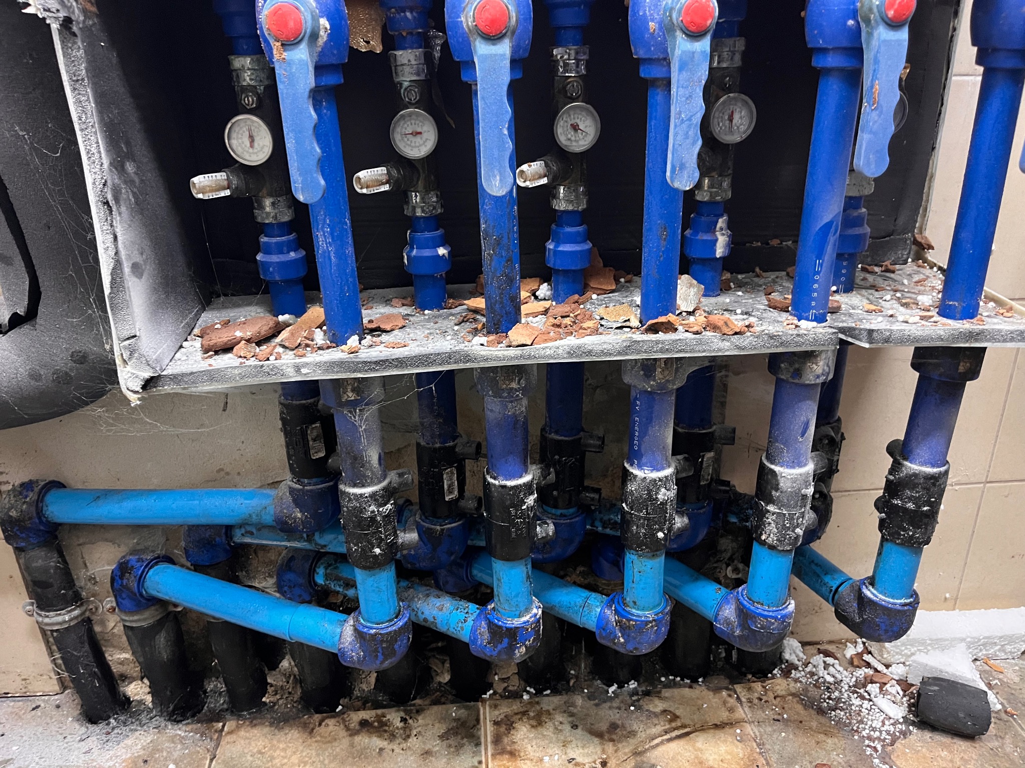 Niebieskie rury instalacji grzewczej z zaworami i manometrami, widoczne połączenia i fragmenty ściany. Instalacja po demontażu, widoczne ślady gruzu i zabrudzenia.