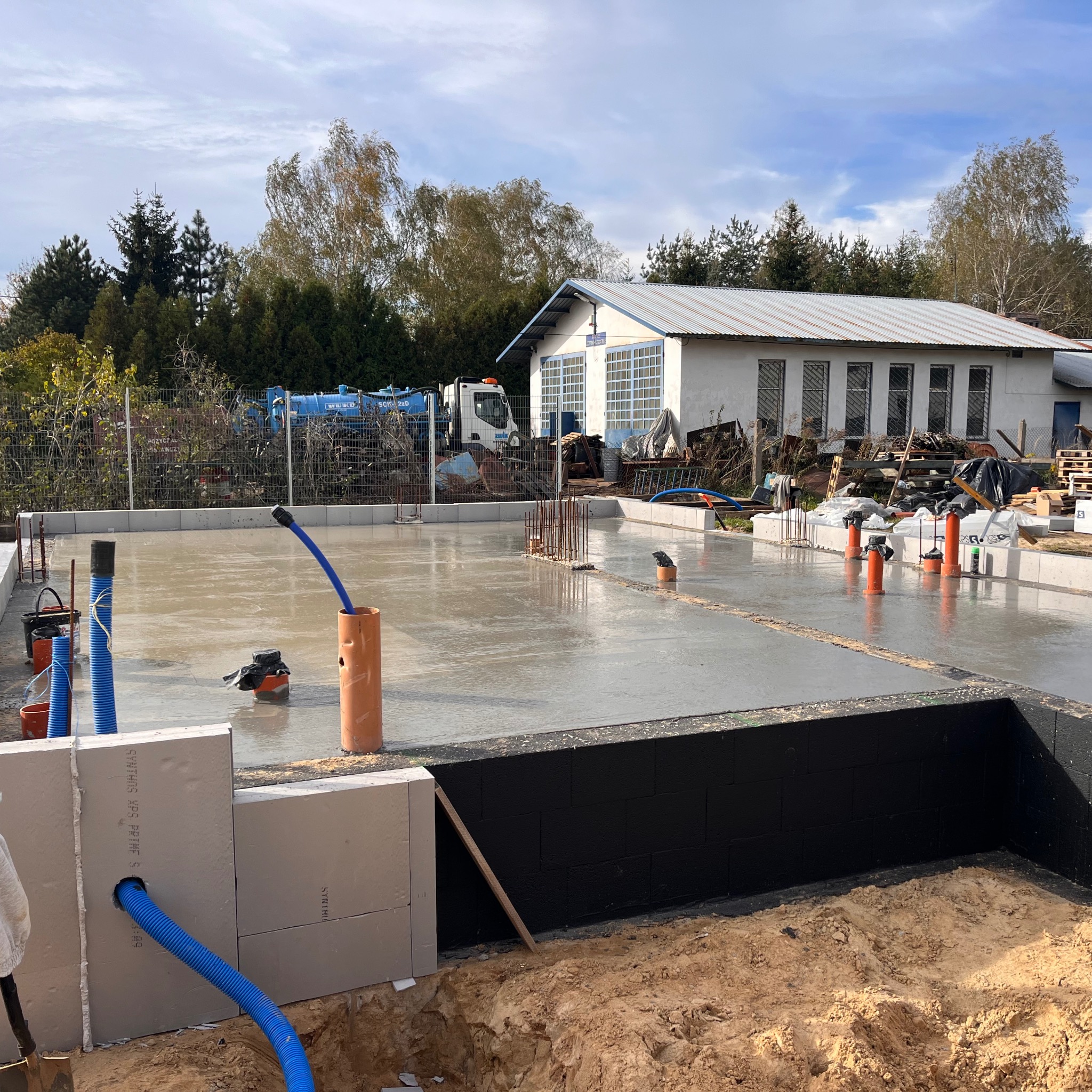 Wylany fundament z instalacją kanalizacyjną i bloczkami izolacyjnymi. Widoczny budynek w tle. Ujęcie z poziomu gruntu, instalacja w kolorze niebieskim i pomarańczowym.