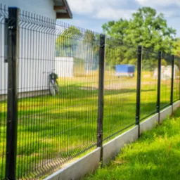 Długi, czarny system ogrodzeniowy z paneli z podmurówką betonową, biegnący wzdłuż trawnika i budynku z rowerem w tle.