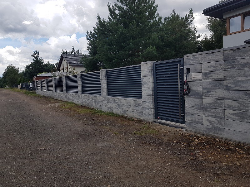 Nowoczesne ogrodzenie z szarych, betonowych bloczków i poziomych paneli, z bramą wjazdową, widok z zewnątrz na tle domów i drzew.