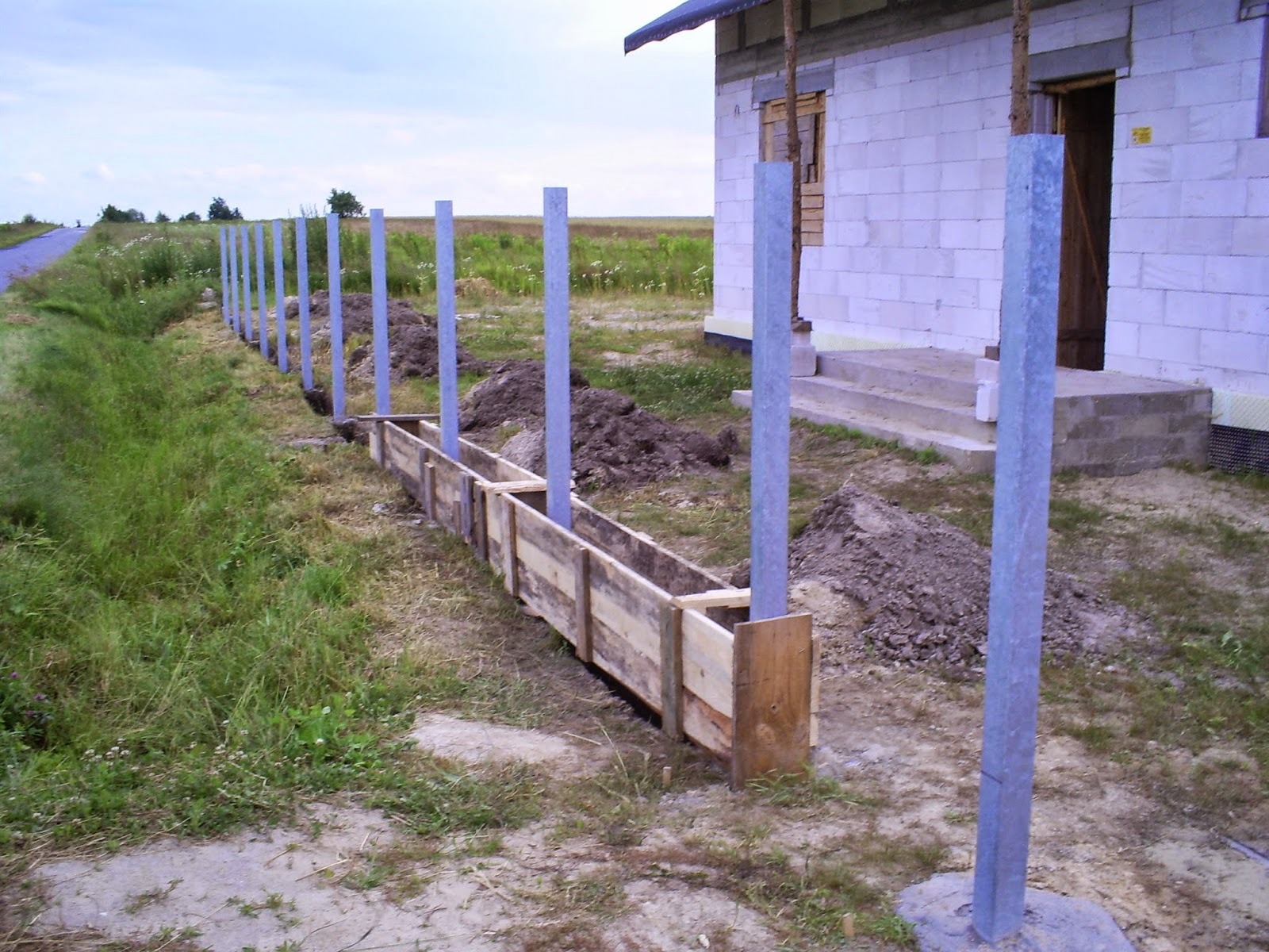 Montaż ogrodzenia betonowego: szalunek drewniany pomiędzy słupkami stalowymi zabetonowanymi w ziemi, w tle budynek w trakcie budowy i zielone pole.